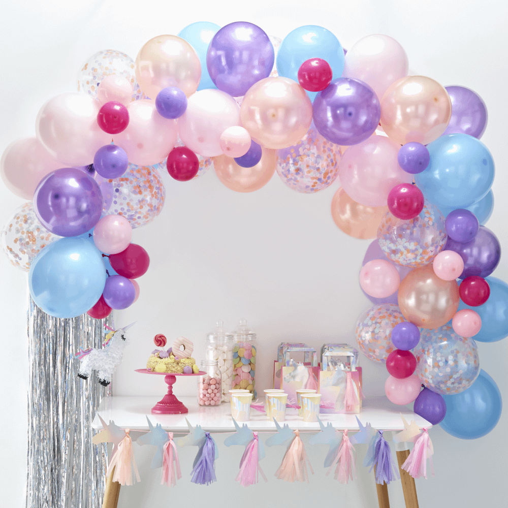 Ballonnenboog met paarse, roze en perzikkleurige ballonnen hangt boven een witte tafel met zilveren tassels en discoballen