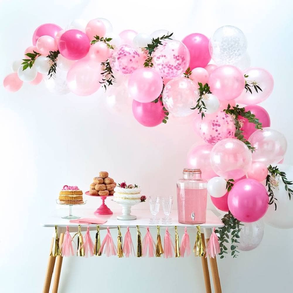 Versierde feesttafel met taart, een tap met drinken en daarboven een roze ballonnenboog met groene takjes