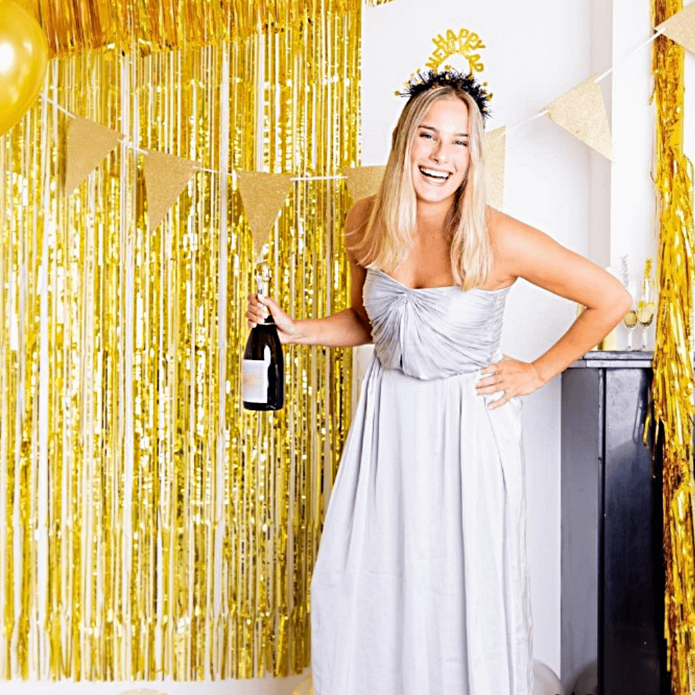 vrouw in witte jurk staat voor een gouden backdrop met een fles champagne