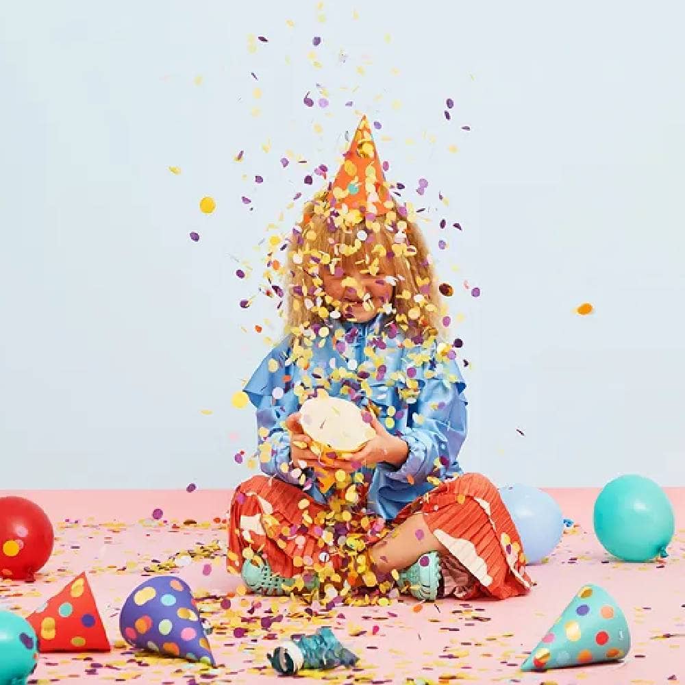 Kind met feesthoedje op en feesthoedjes om zich heen speelt met confetti