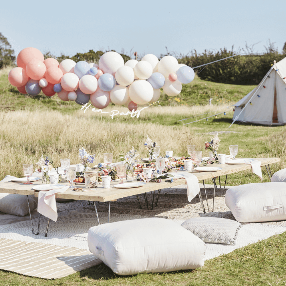 Picknicktafel versierd met bloemen staat in een grasveld met witte kussens en een pastelkleurige, matte ballonboog