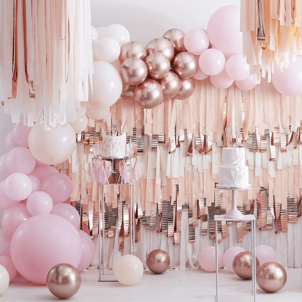 Rosé gouden met roze ballonboog en reflecterende streamers hangt aan een muur achter twee wit met roze taarten