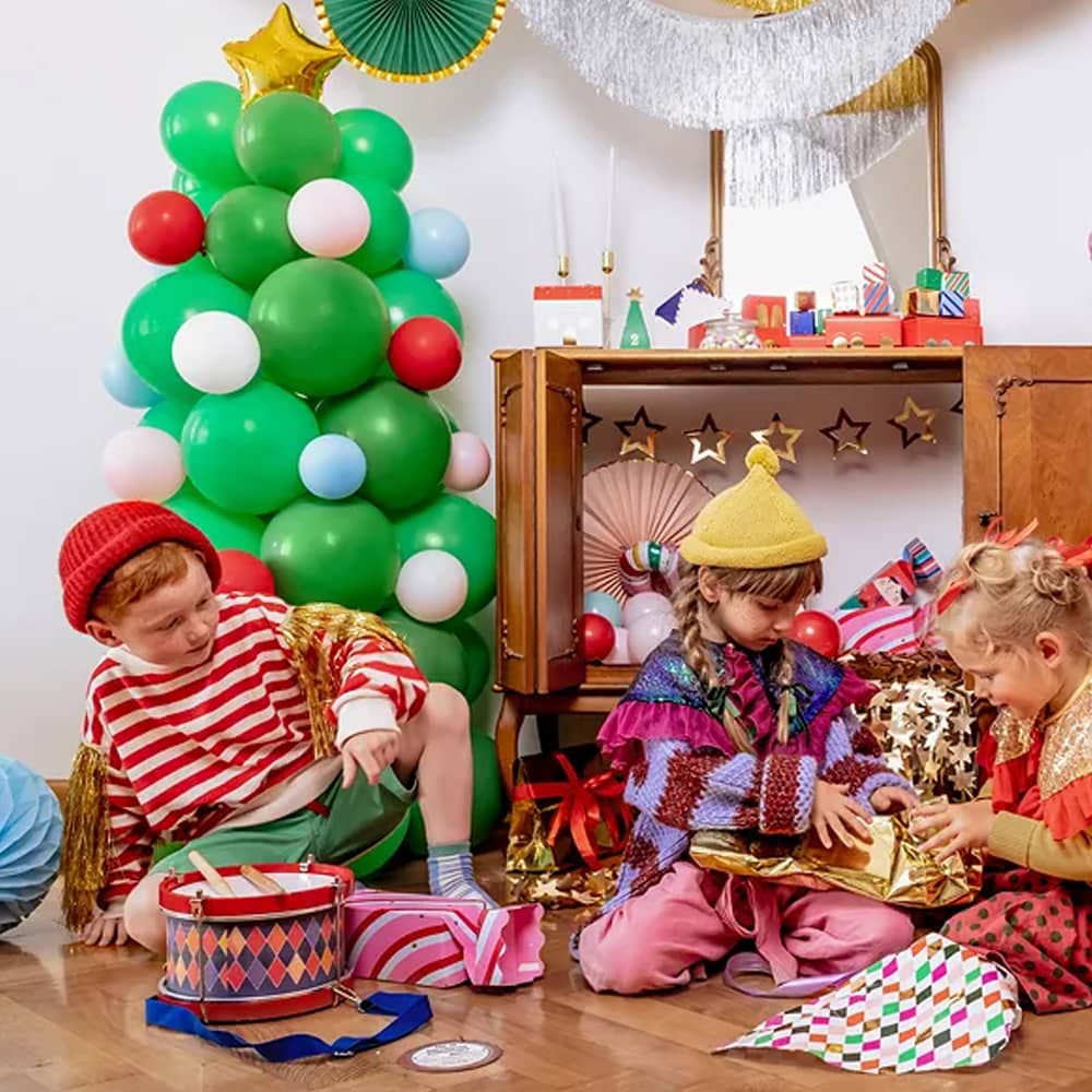 Drie kinderen in een versierde kamer met slingers waaiers en een kerstboom van ballonnen