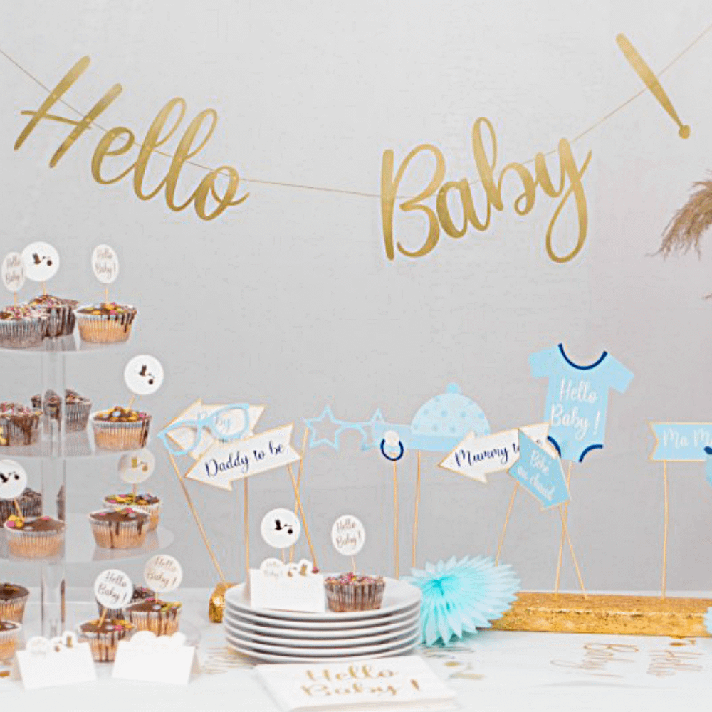 Gouden hello baby slinger hangt voor een grijze muur boven een tafel met versiering voor een babyshower
