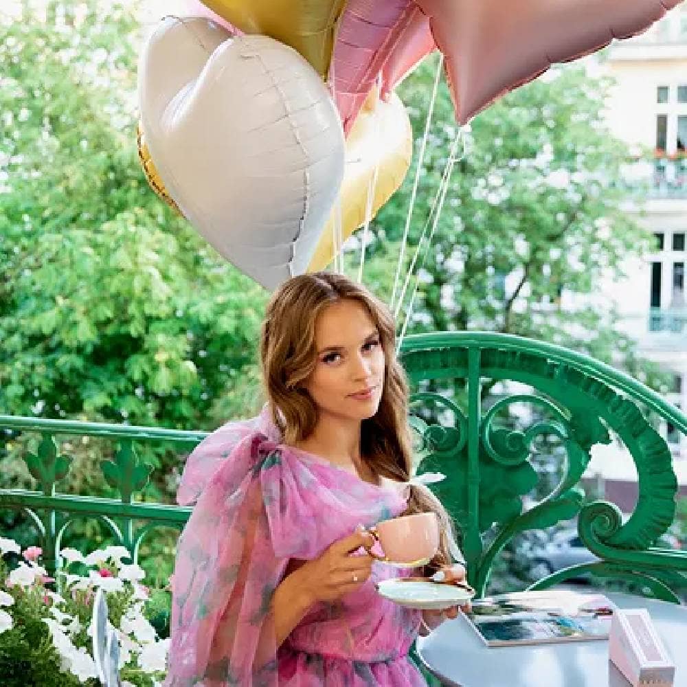 Vrouw met een roze kopje op een balkon zit voor een ballon boeket met hartvormige ballonnen