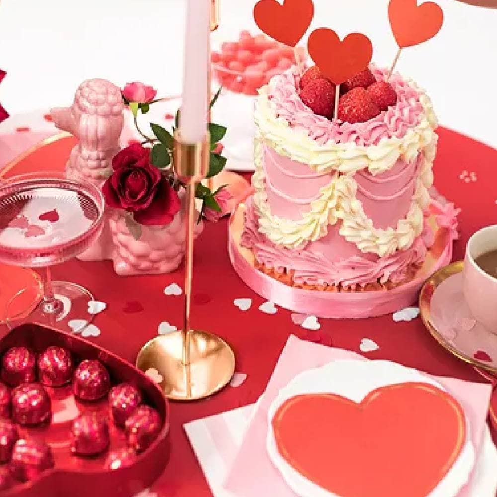 Tafel met rood kleed, rode hartvormige servetten, een roze taart en witte, hartvormige confetti