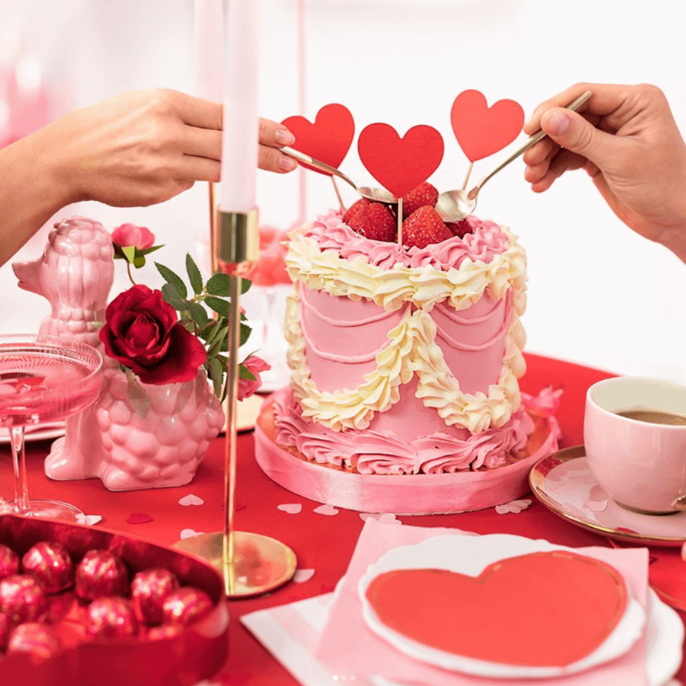 Roze taart versierd met rode hartjes staat op een tafel met rood tafelkleed voor een gouden kaarsenhouder