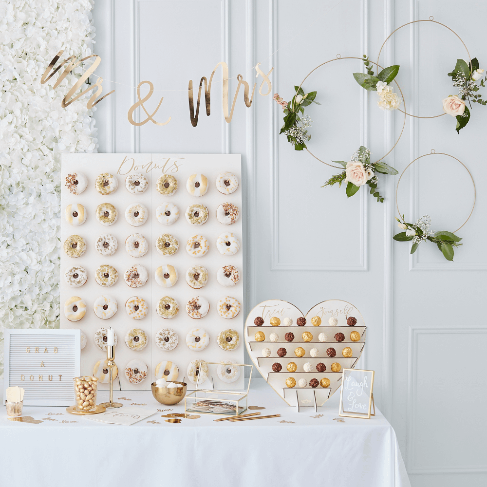 Witte tafel staat voor een witte muur en is bedekt met een donut wall, een hartje, een fotolijstje en gouden versiering