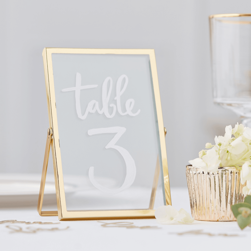 Gouden fotolijst met glazen plaatjes waar een witte tekst op is geschreven staat op een tafel versierd met confetti en witte bloemblaadjes