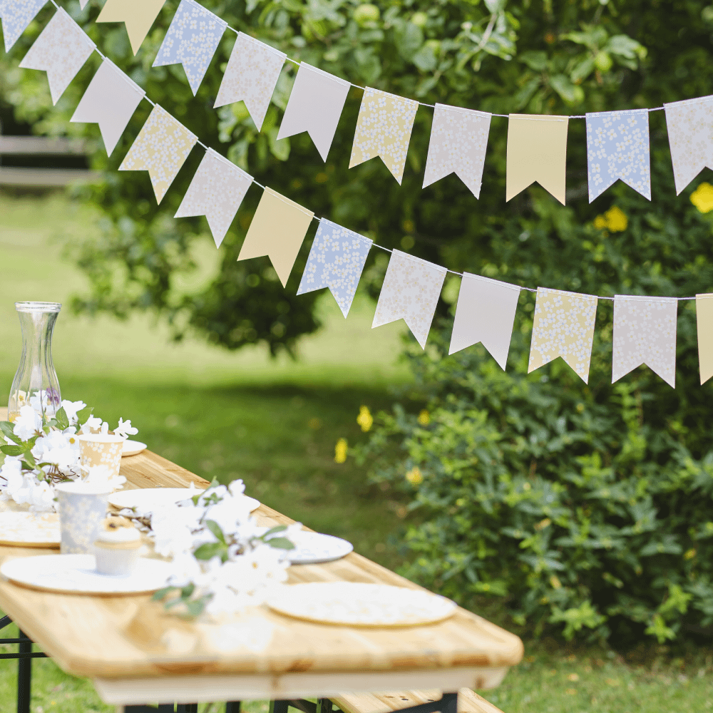 Pastelslingers met bloemenprint hangen boven een houten picknicktafel versierd met bloemen en een glazen vaas