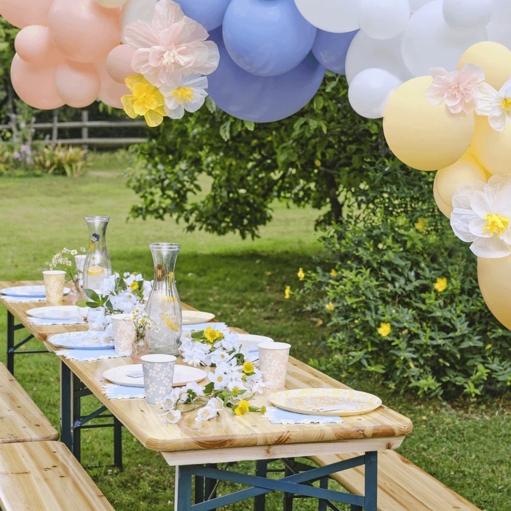 Picknicktafel staat op een grasveld en is versierd met bloemen en papieren bordjes en bekers in pasteltinten