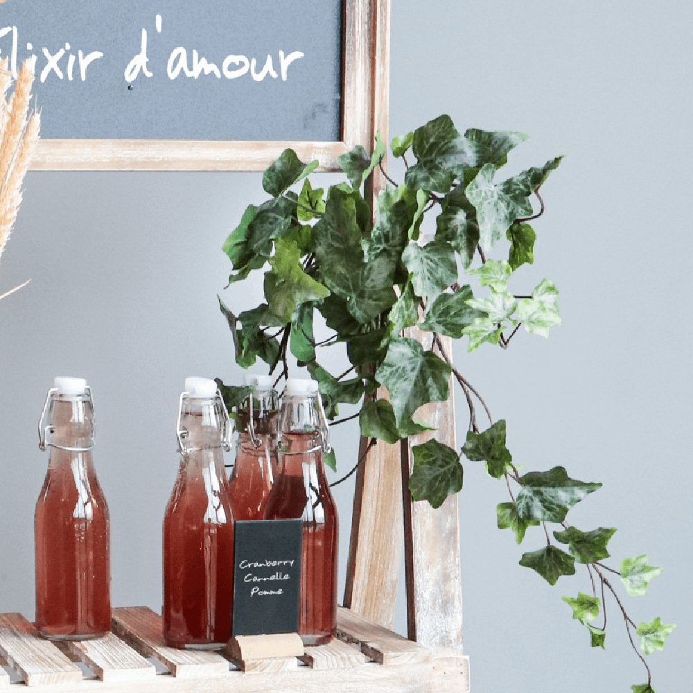 Houten kraam met een plant eraan is voorzien van glazen flessen met een rode drank en een krijtbord met Franse tekst