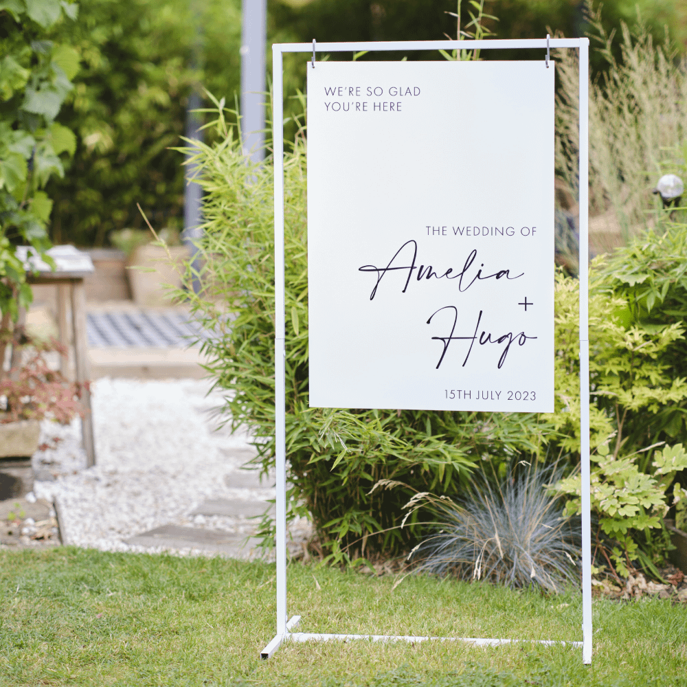 Witte metalen standaard staat in een grasveld en in het midden hangt een bord met een uitnodiging voor een bruiloft