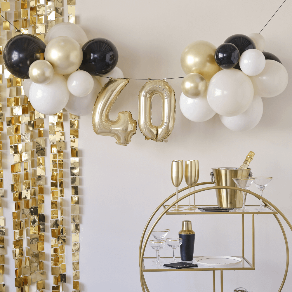 Slinger voor 40 jaar met gouden, nude en zwarte ballonnen hangt voor een backdrop met gouden vierkantjes en boven een rond gouden karretjes met een emmer met champagne en een cocktailshaker