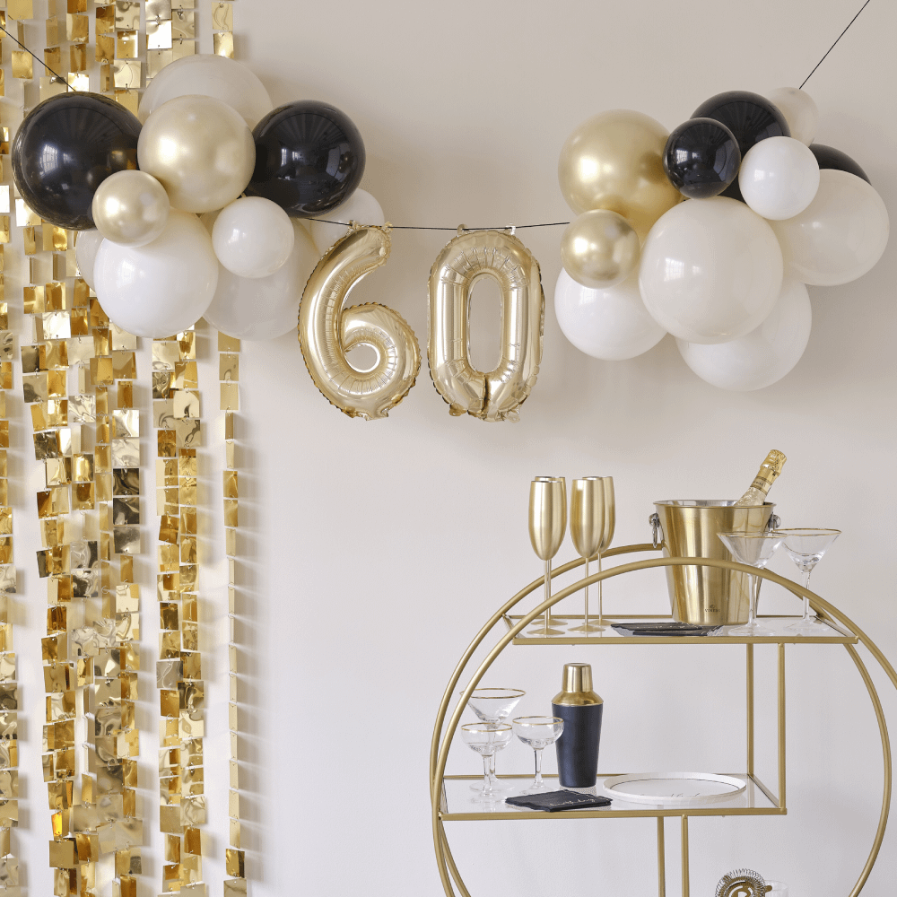 Ballonnenslinger voor 60 jaar in het goud, nude en zwart hangt boven een gouden karretje met champagneglazen en een cocktailshaker en voor een gouden backdrop met vierkantjes
