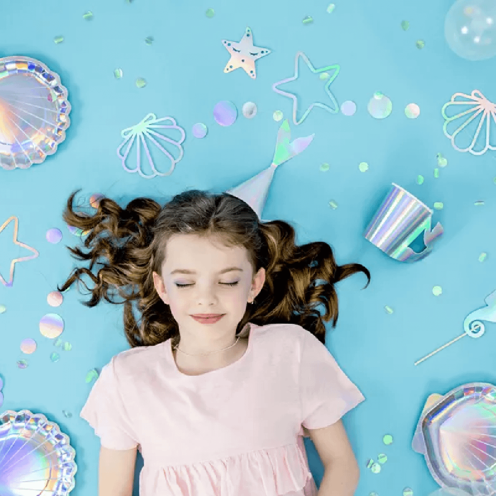 Meisje met bruin haar ligt op een blauwe ondergrond en is omringd met iridescent versiering van zeesterren, schelpen en zeemeerminnen