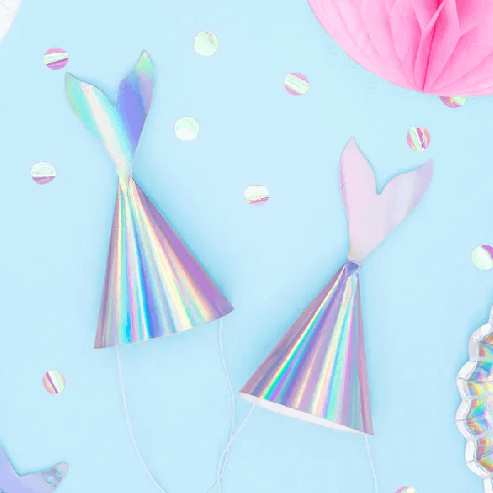 Irirdescent feesthoedjes met zeemeerminnenstaart liggen op een lichtblauwe ondergrond met ronde confetti