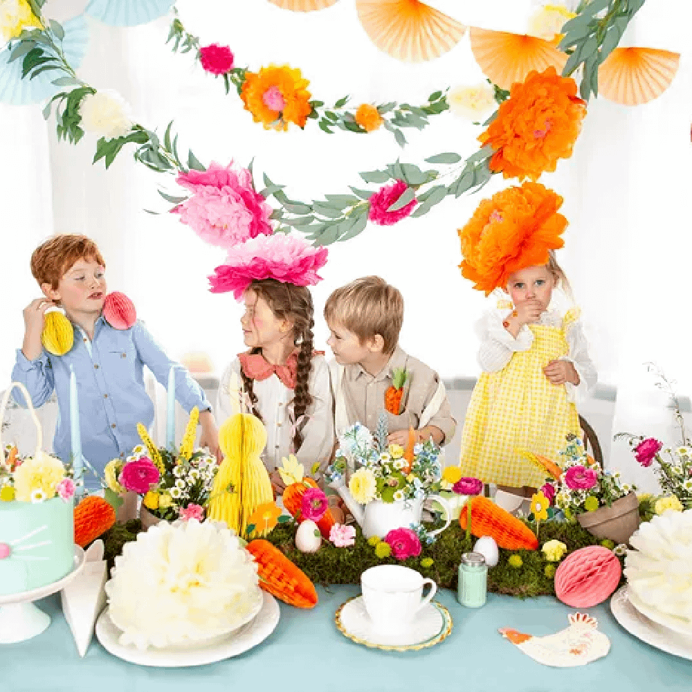 Vier kinderen zitten aan een tafel met lichtblauw tafelkleed versierd met papieren bloemen en honeycombs in de vorm van paaseieren