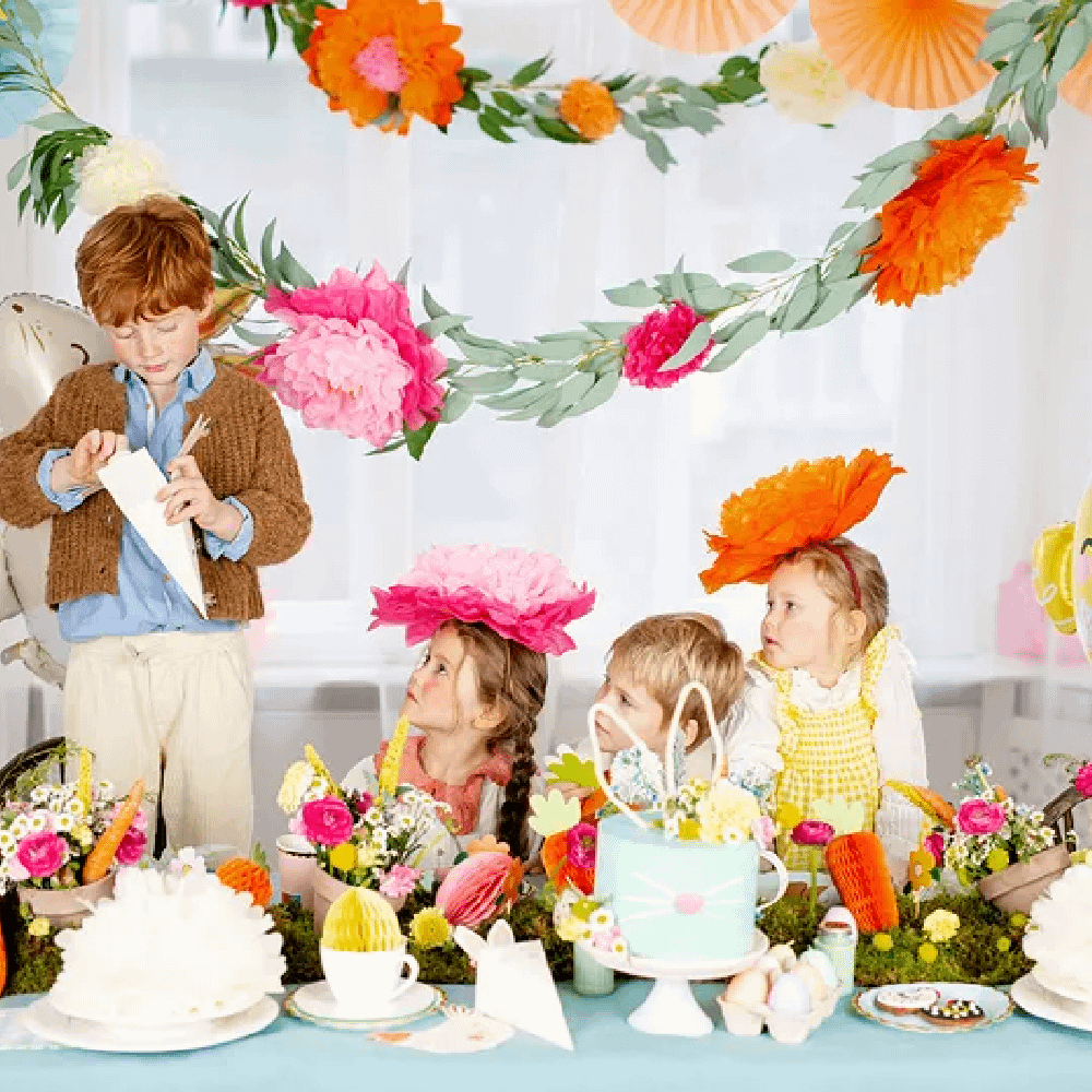 Drie kinderen zitten aan een tafel met lichtblauw tafelkleed versierd met papieren bloemen en honeycombs in de vorm van paaseieren