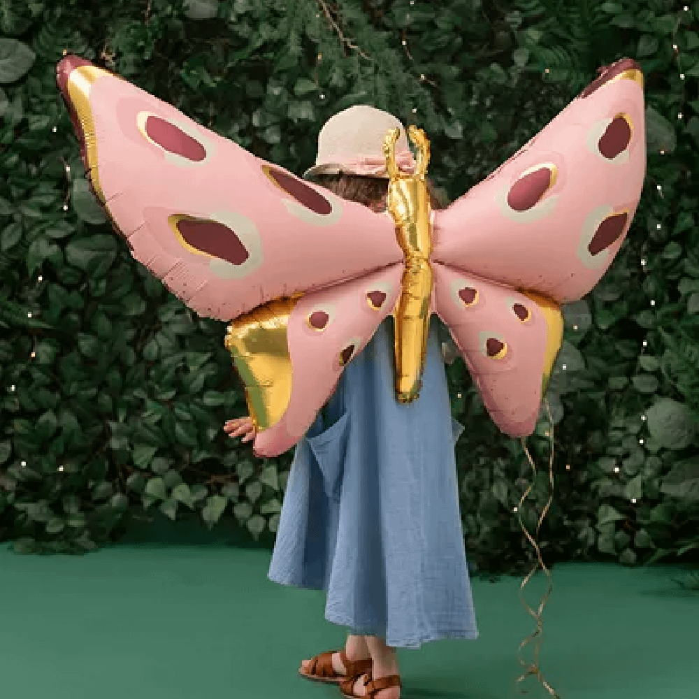 Meisje met blauwe jurk en bruin haar staat voor een heg en heeft een folieballon vast in de vorm van een vlinder in het roze, goud en paars