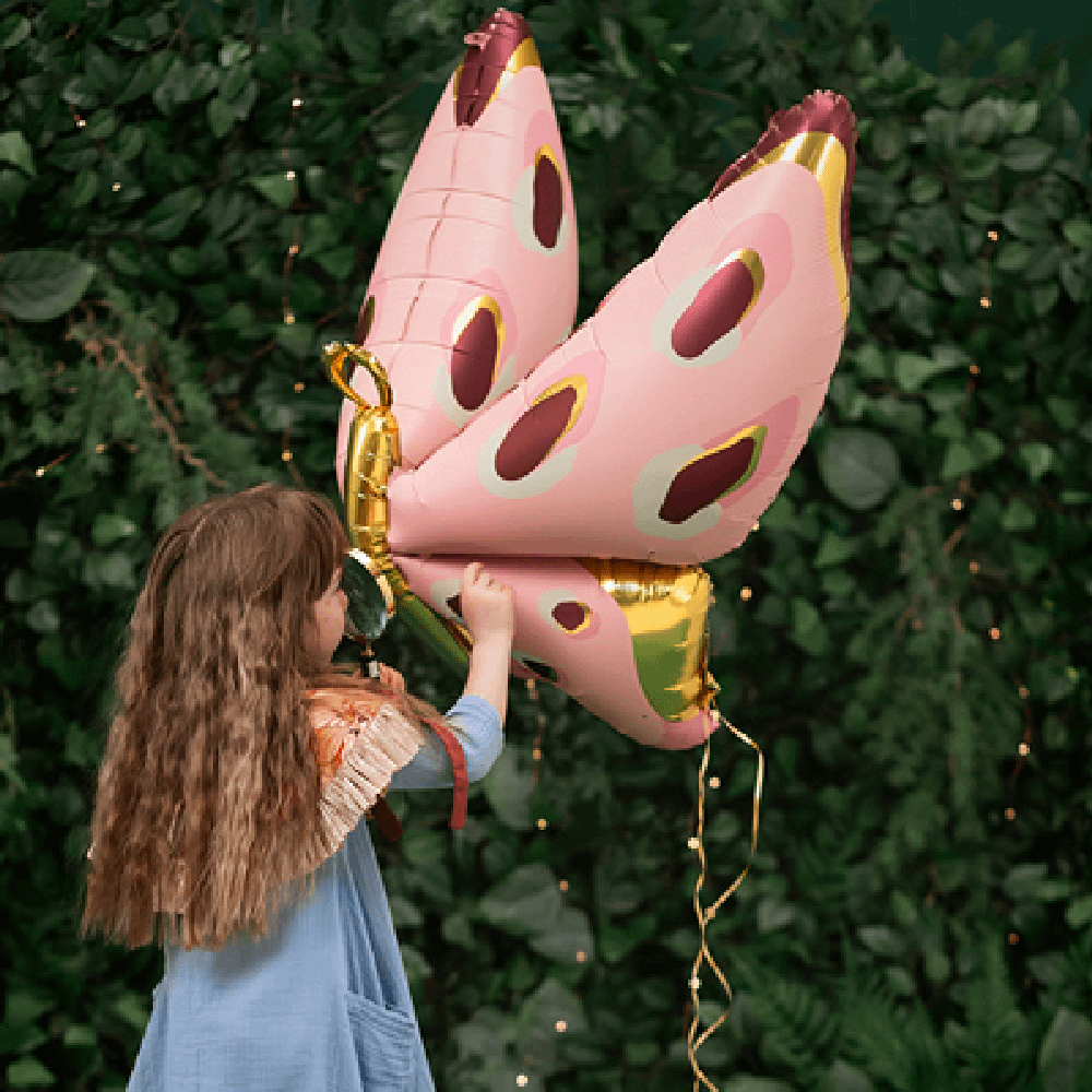 Meisje met blauwe jurk en bruin haar staat voor een heg en heeft een folieballon vast in de vorm van een vlinder in het roze, goud en paars