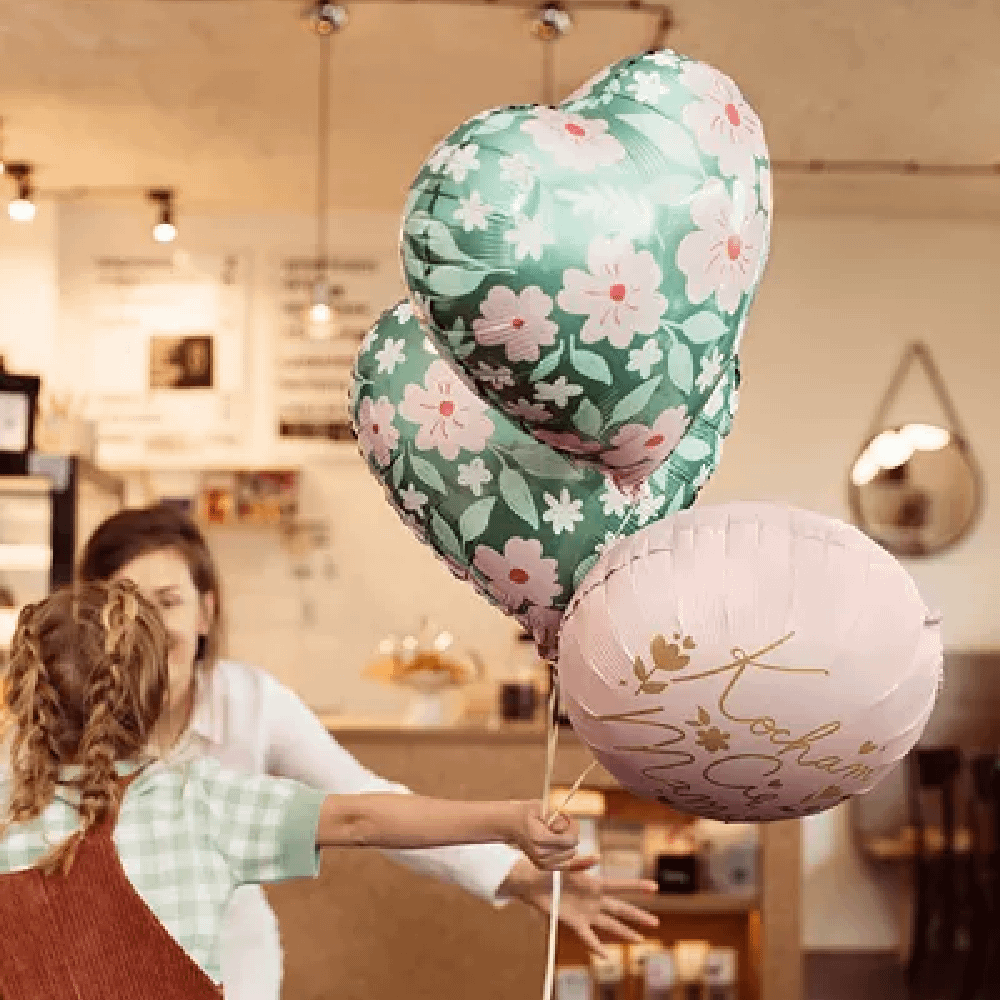 Meisje met vlechtjes houd een ballonnenboeket vast met een groene ballon in de vorm van een hart en een roze ballon met gouden tekst