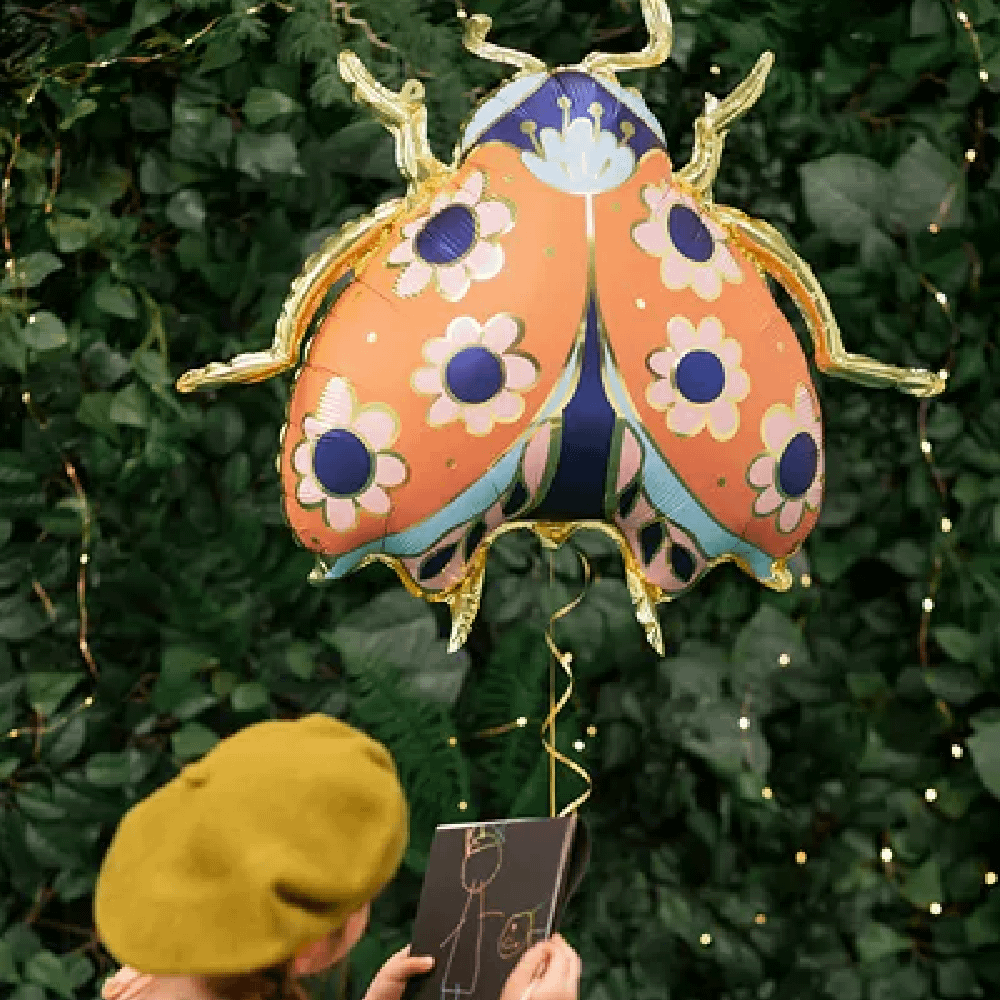 Meisje met geel hoedje staat voor een hef met een folieballon in de vorm van een lieveheerstbeestje met gouden poten