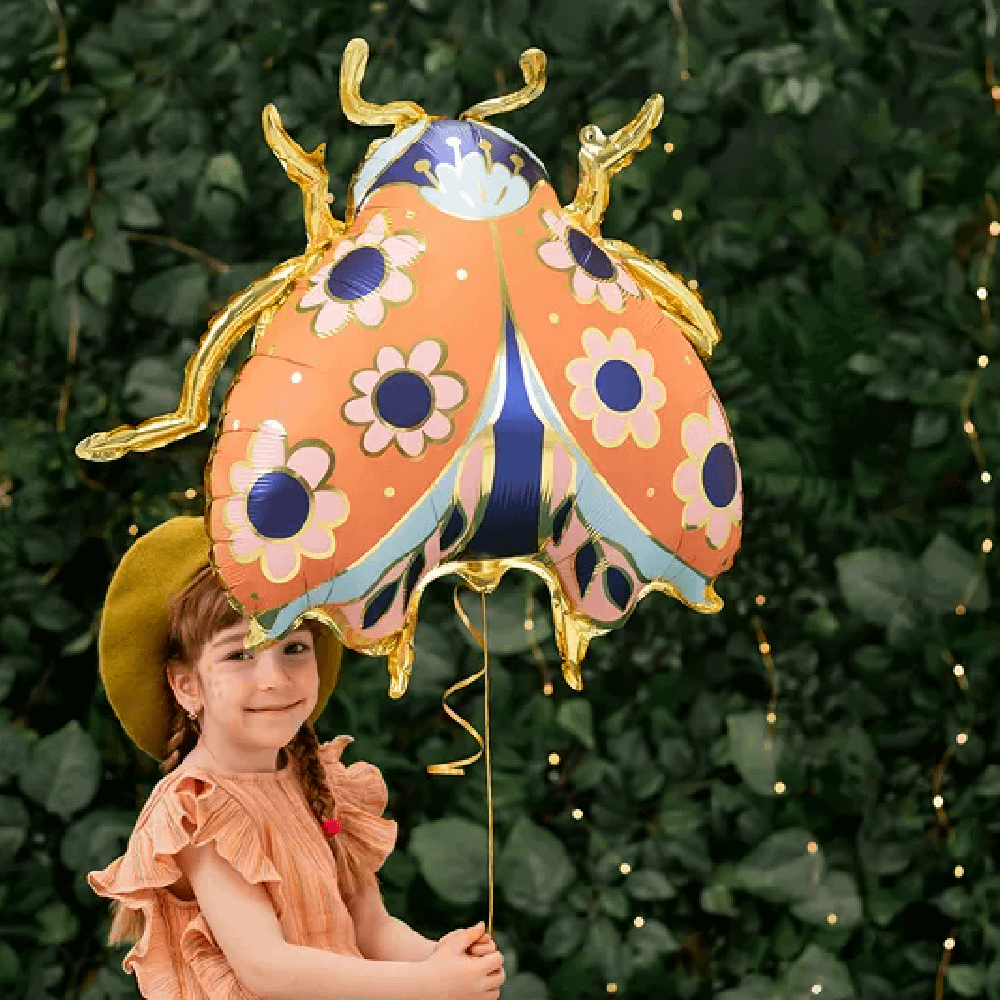 Meisje met geel hoedje staat voor een hef met een folieballon in de vorm van een lieveheerstbeestje met gouden poten
