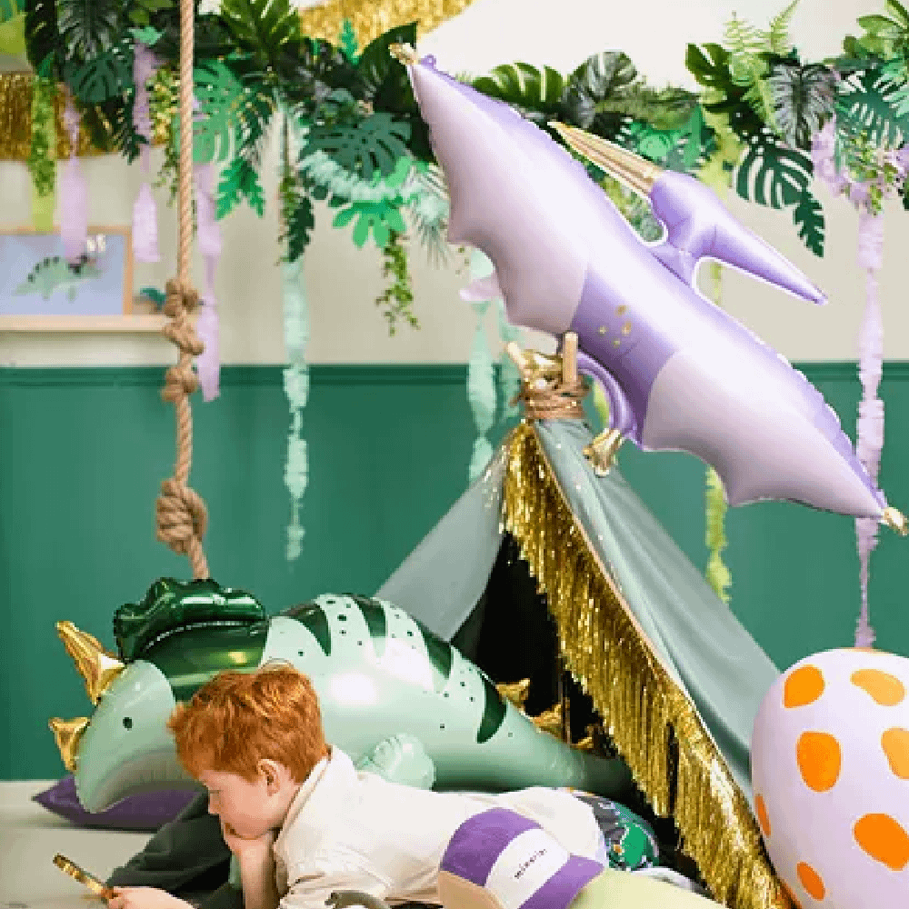 Jongetje ligt in een groene tent met gouden franjes in een kamer versierd met palmbladeren en folieballonnen in de vorm van dino's