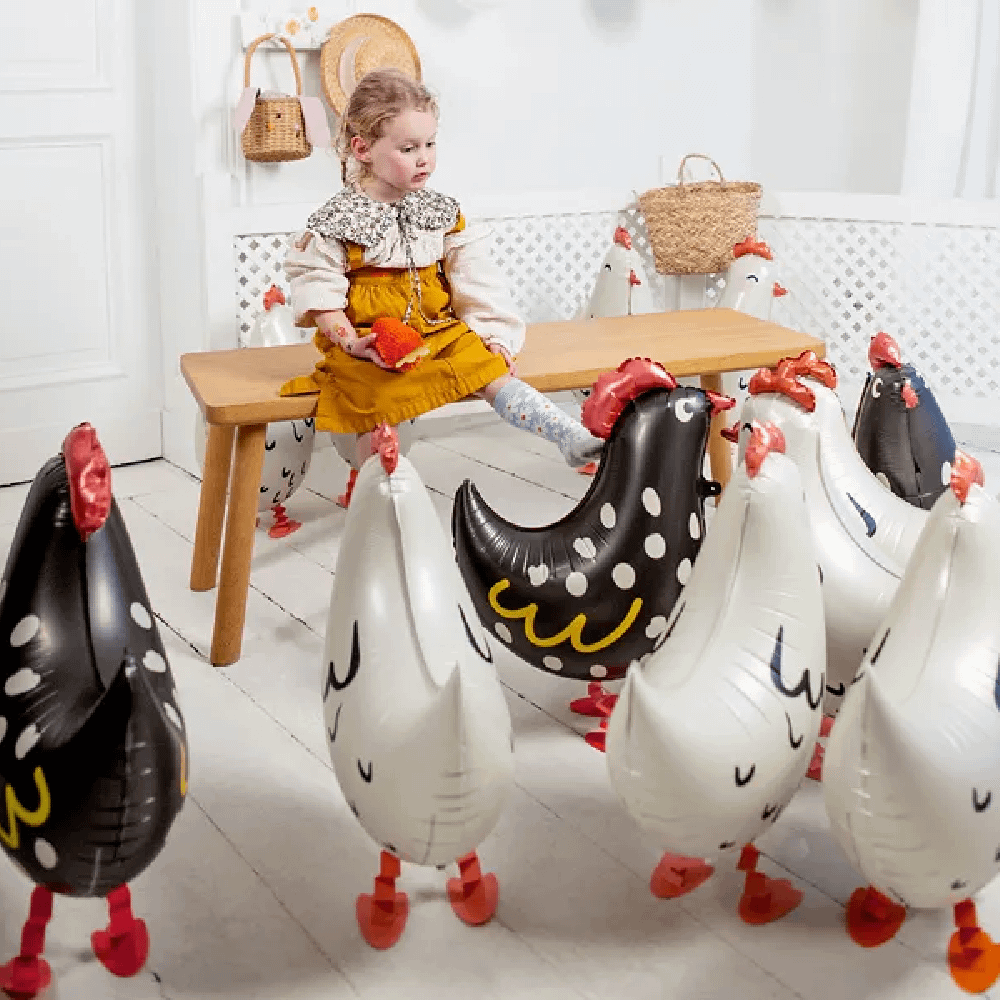 Meisje zit op een houten bankje in een kamer met folieballonnen in de vorm van kippen in het wit en zwart