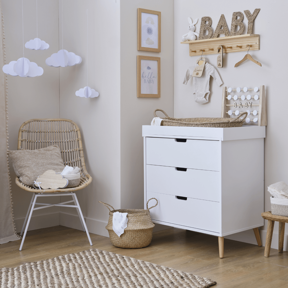 Babykamer met een houten vloer, beige muur en witte kast versierd met houten kledinghangers, een houten plank, een rieten stoel en papieren wolken