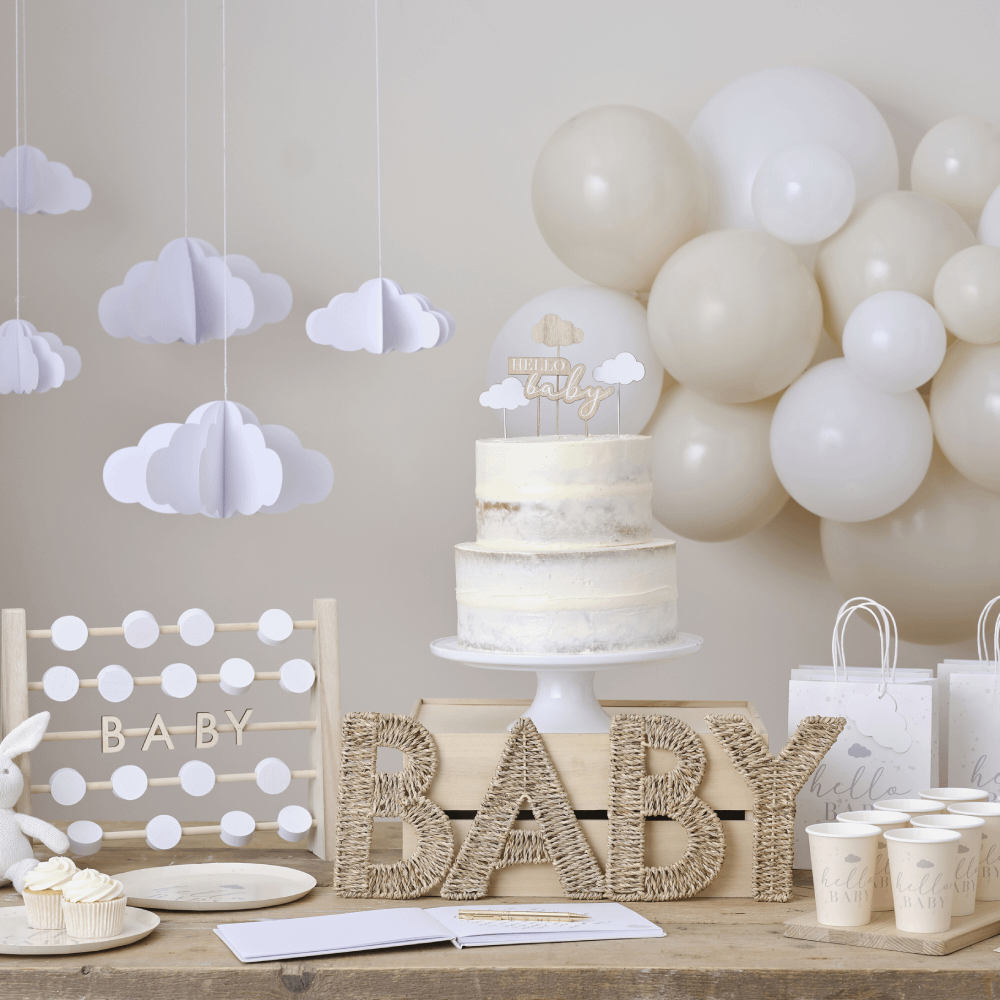 Houten tafel staat voor een beige muur versierd met papieren wolkjes en beige ballonnen, een rieten bord met de tekst 'baby' en een witte taart met houten toppers