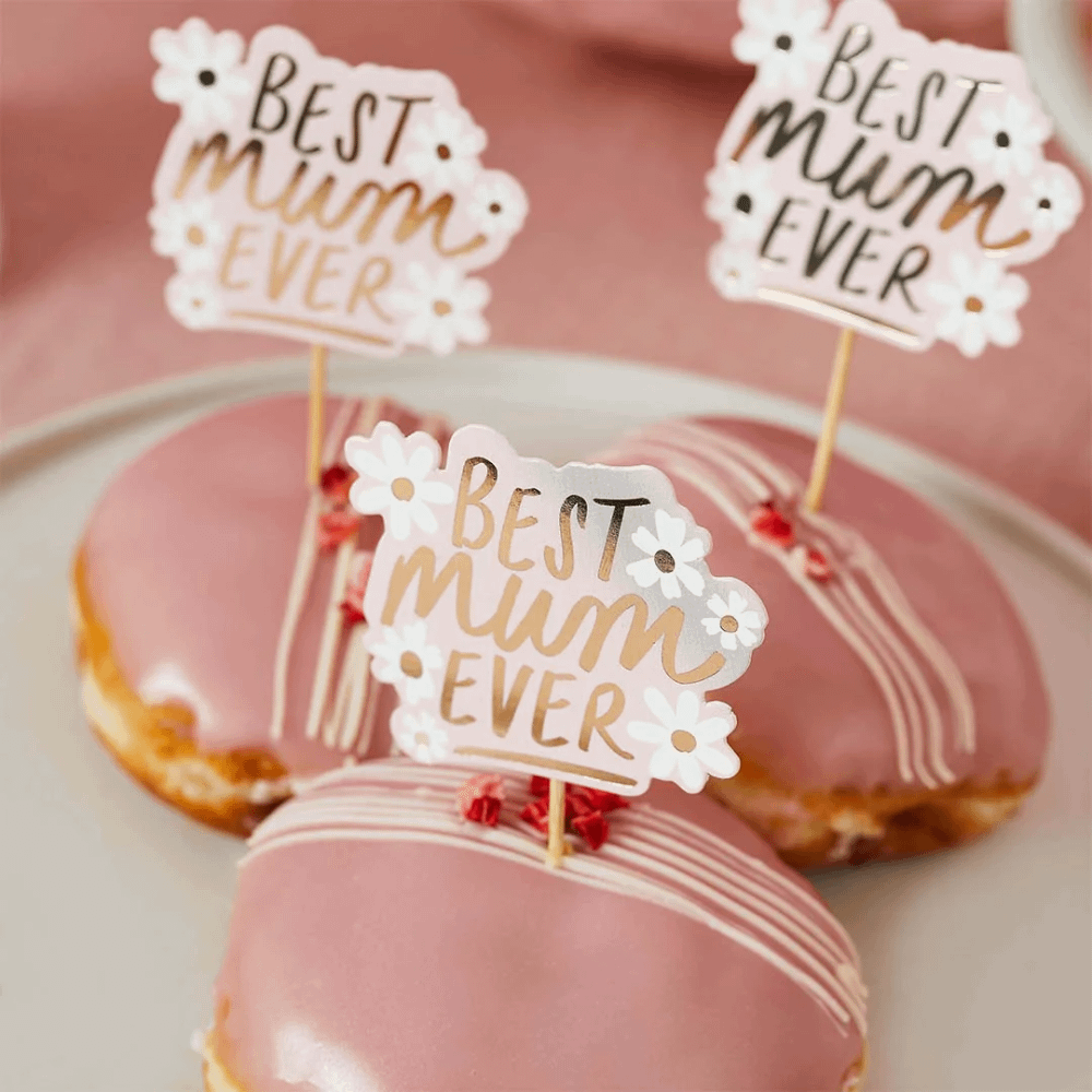 Koeken met roze laag liggen op een wit bord en zijn versierd met cupcakeprikkers met madeliefjes en de tekst best mum ever