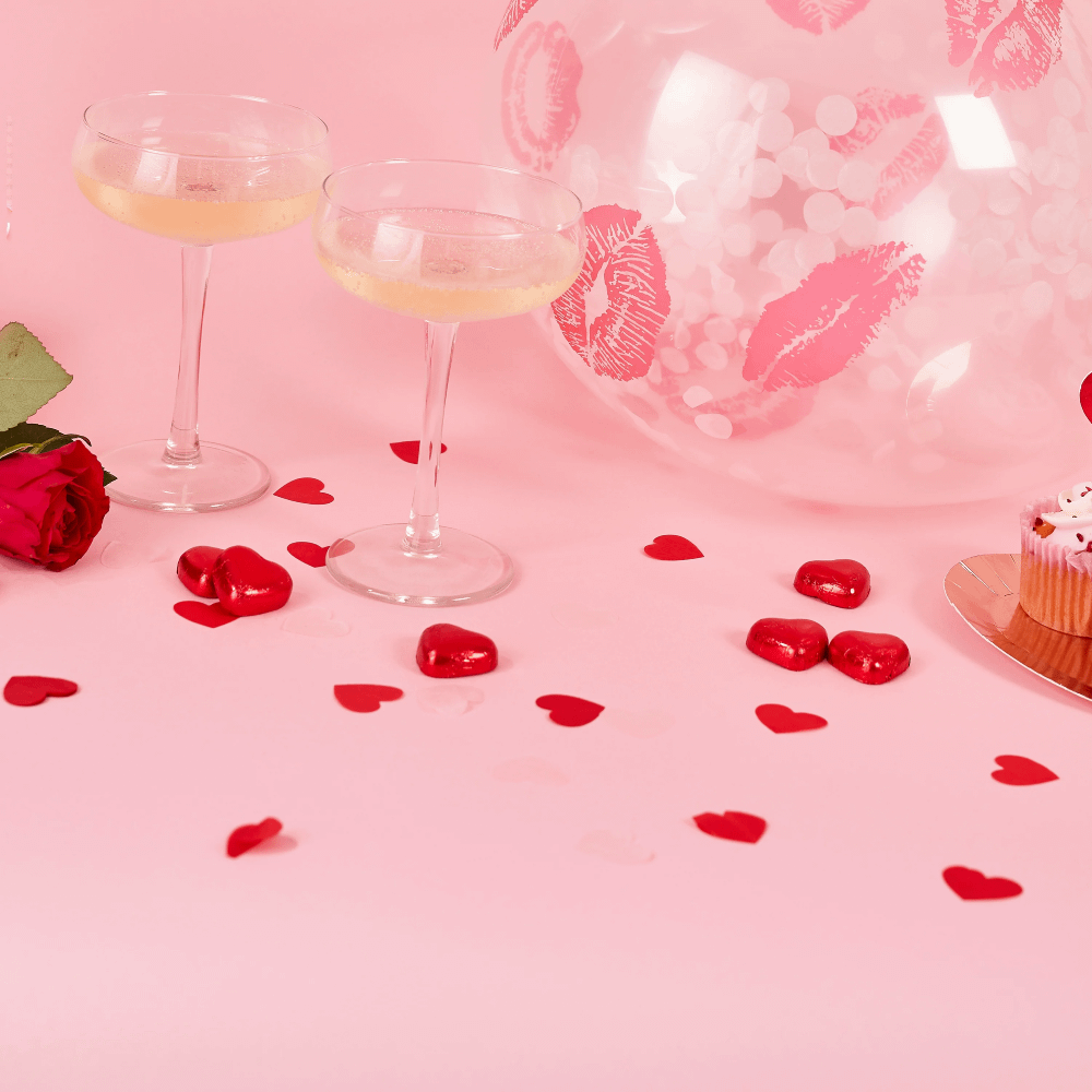 Roze tafel met glazen met champagne, hartvormige confetti en een transparante ballon met roze lippen erop