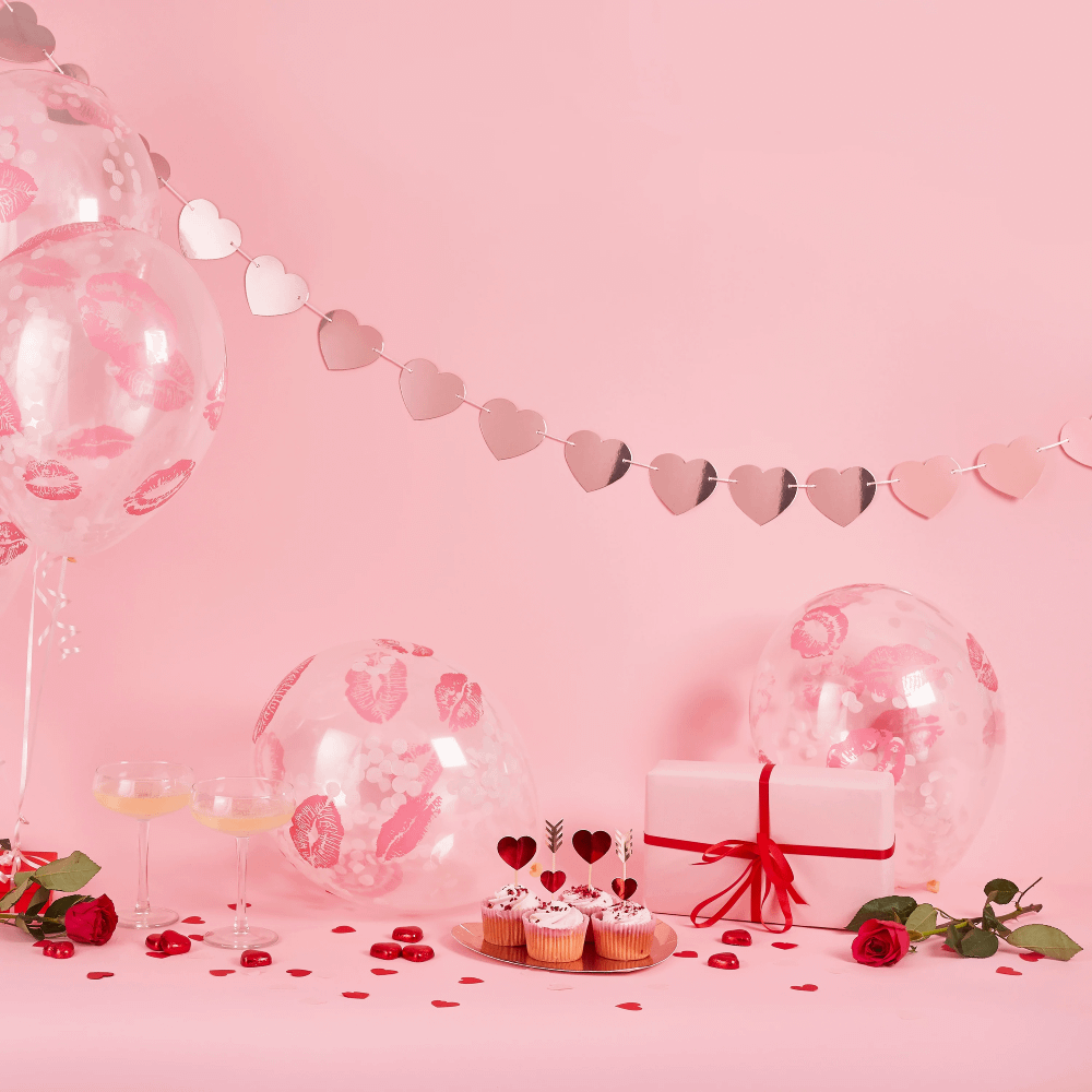 Rose gouden hartjesslinger hangt voor een roze achtergrond en boven transparante ballonnen, een bord met cupcakes en een rode roos