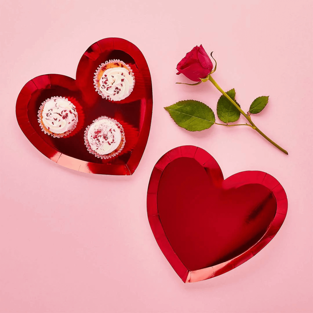 Papieren bordjes met metallic afwerking in de vorm van een hart liggen op een roze ondergrond naast een rode roos en zijn gevuld met cupcakes