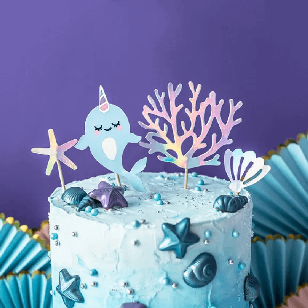 Blauw met witte taart versierd met schelpen en cupcaketoppers in de vorm van een narwal, zeewier, een zeester en een schelp met iridescent effect