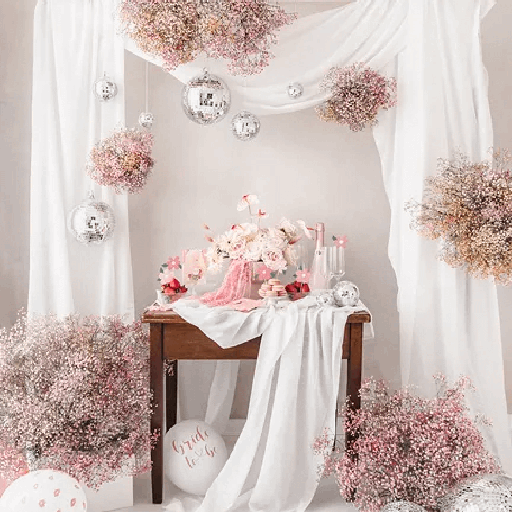 Bruine tafel versierd met een wit kleed en witte bloemen staat tussen roze planten en wit met roze ballonnen