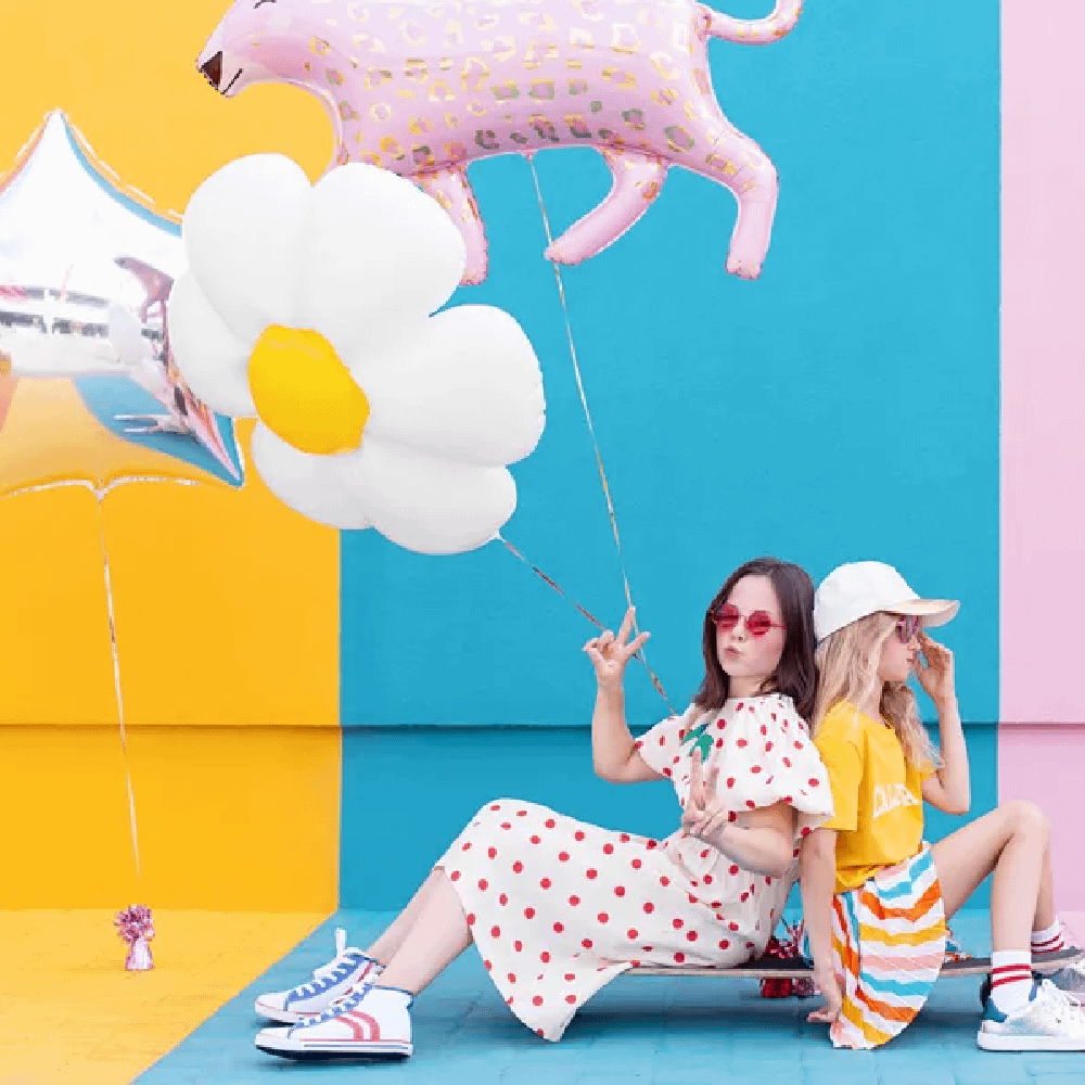 Twee meisjes zitten op een skateboard en houden folieballonnen vast in de vorm van een madeliefje, een roze tijger en een zilveren ster