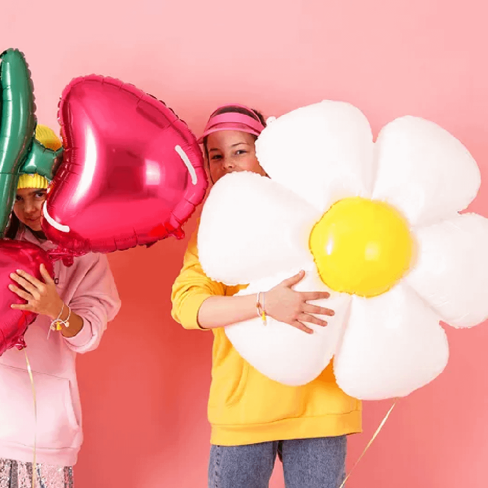 Meisje in gele trui houdt een folieballon vast in de vorm van een madeliefje en staat voor een roze achtergrond