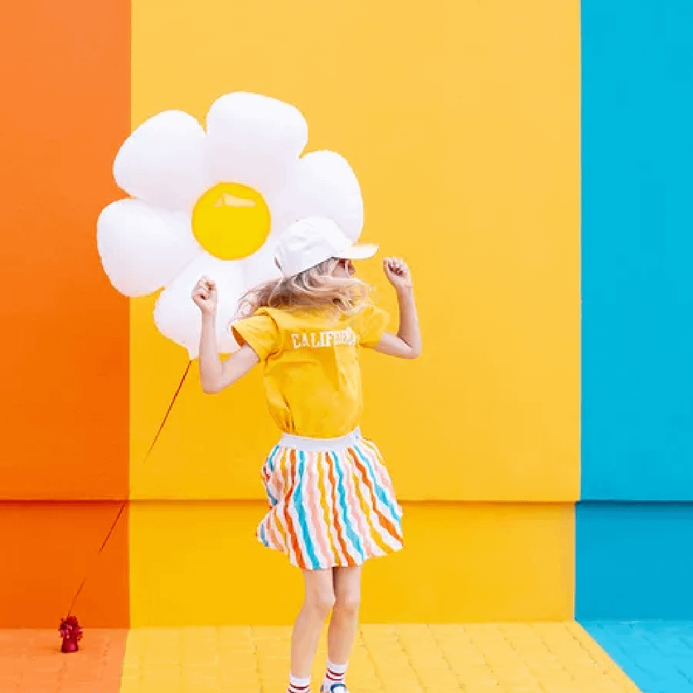 Meisje met geel shirt houdt een ballon vast van een madeliefje en staat voor een oranje, gele en blauwe achtergrond