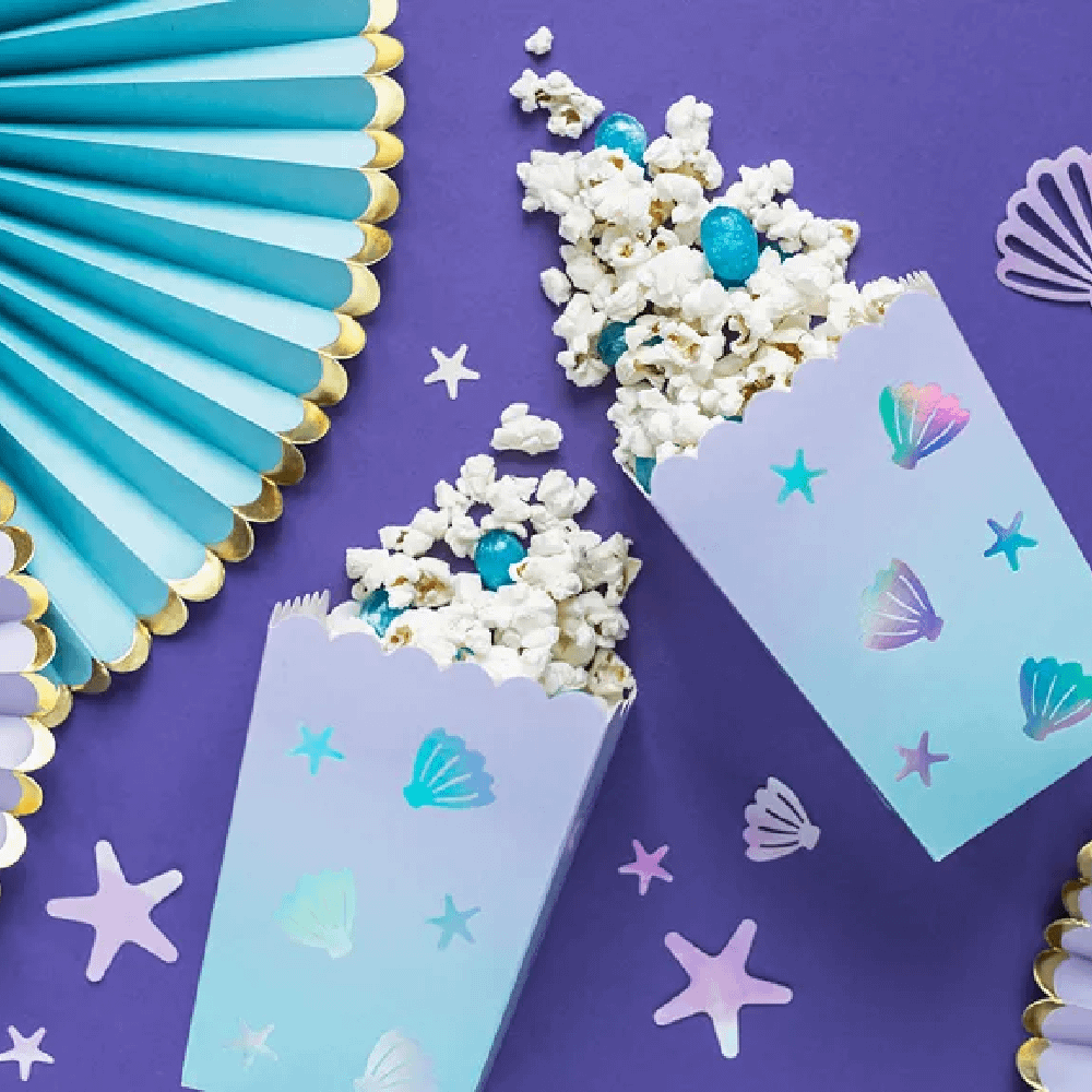 Popcornbakjes gevuld met popcorn liggen op een paarse tafel versierd met iridescent confetti en een lichtblauwe waaier met gouden rand