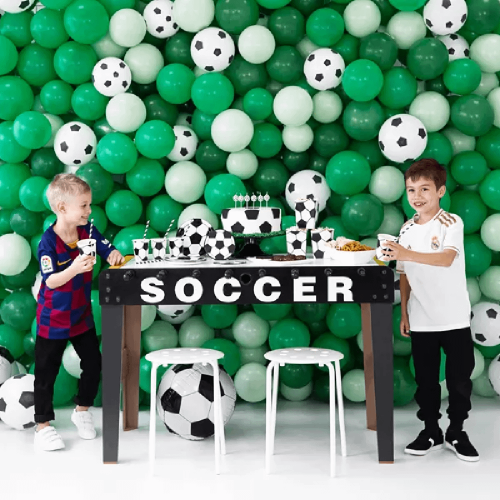 Twee jongens staan bij een voetbaltafel voor een muur bedekt met groene en witte ballonnen met voetbalprint