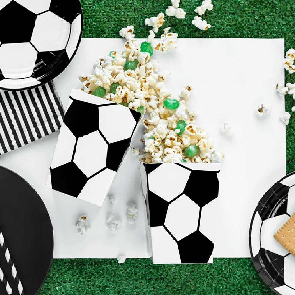 Kartonnen bakjes, rietjes en bordjes met voetbalprint liggen op een grasveld met popcorn