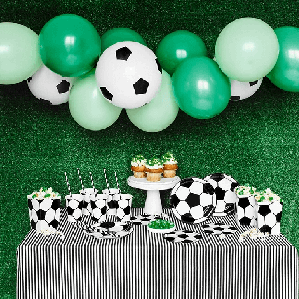 Groene muur van kunstgras is versierd met een ballonnenboog met groene ballonnen en een tafel met bekers, bakjes en bordjes met voetbalprint