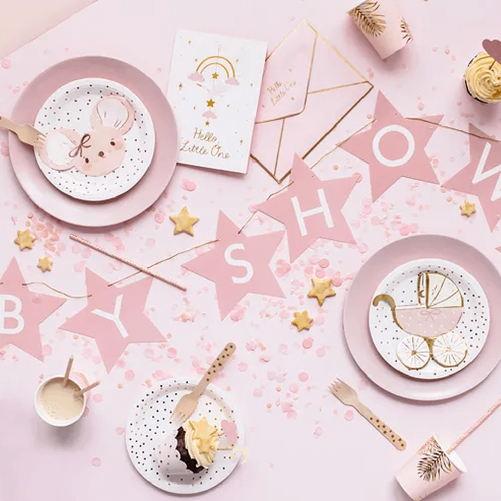 Roze tafel met een roze sterrenslinger met de tekst babyshower, roze bordjes en servetten en papieren bekertjes met gouden palmbladeren