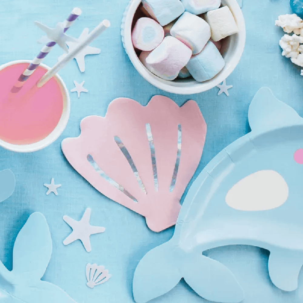 Lichtroze servet in de vorm van een schelp liggen op een blauwe tafel met marshmallows en een beker met roze drinken