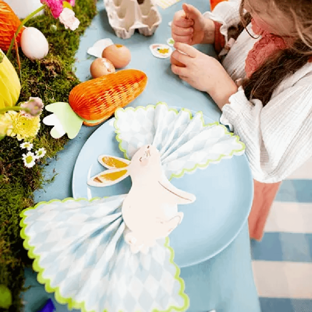 Meisje staat aan een tafel met lichtblauw tafelkleed aangekleed met honeycomb paaseieren en wortels en versierd een paasei