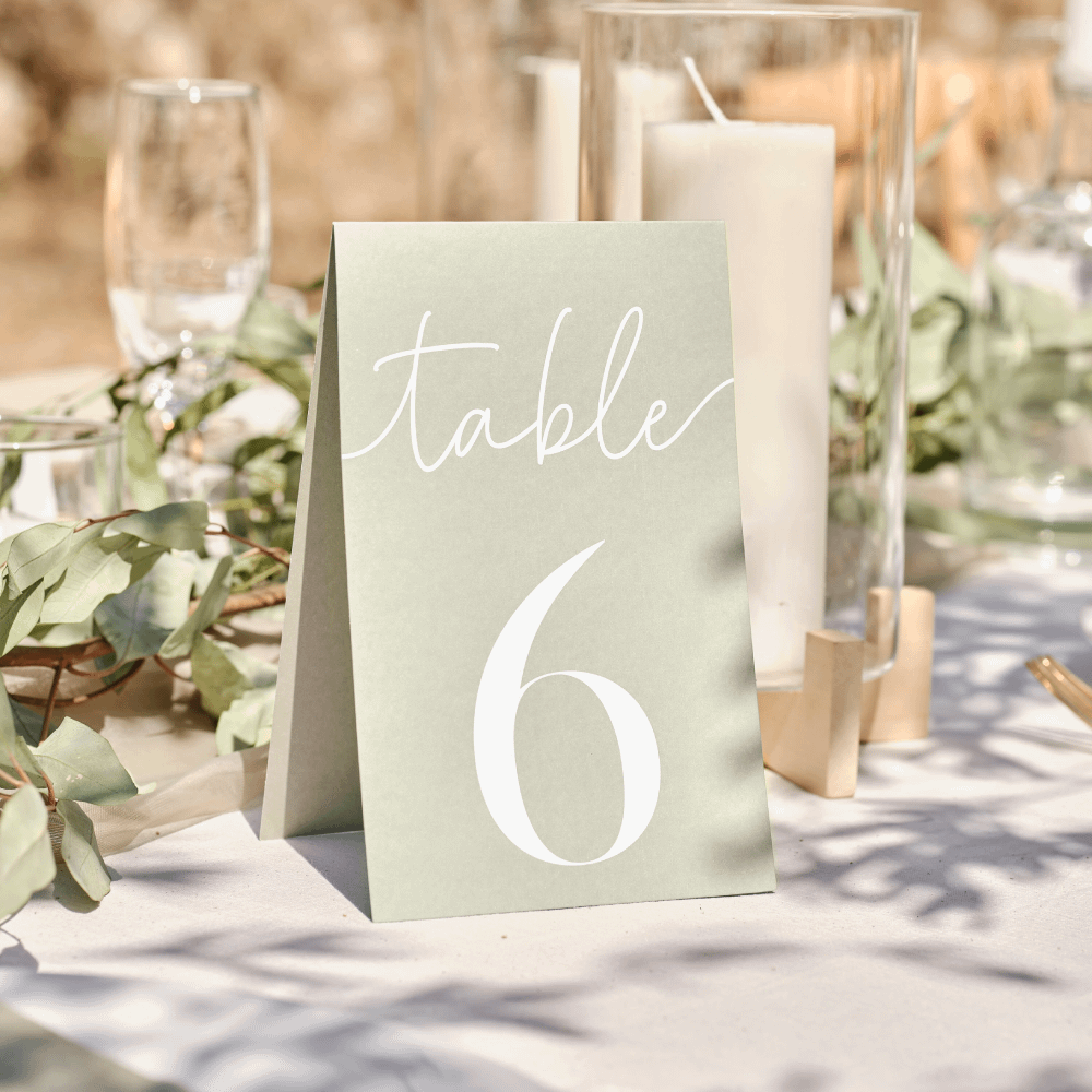 Saliegroen tafelkaartje met witte tekst staat op een witte tafel voor een kaars in glazen houder en wat bladgroen