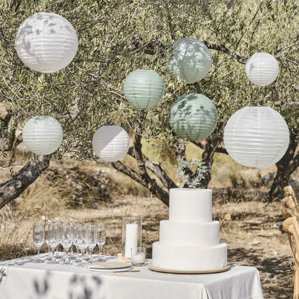 Witte en saliegroene lampionnen hangen aan bomen boven een witte tafel met een taart en champagneglazen